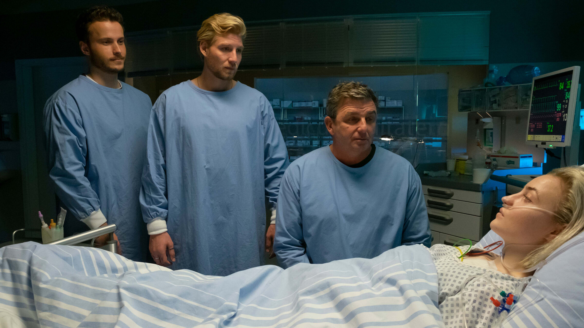 "Der Bergdoktor: Über den Schatten": Im Krankenhaus: Karla Jenner (Sarah Mahita) liegt, angeschlossen an Infusionen und einem Überwachungsmonitor, im Bett. Daneben sitzt Dr. Martin Gruber (Hans Sigl). Neben ihm stehen Markus Jenner (Nicolas Wolf) und Fabian Marx (Hannes Schmid). Alle drei tragen blaue Schutzkittel und blicken Karla besorgt an.