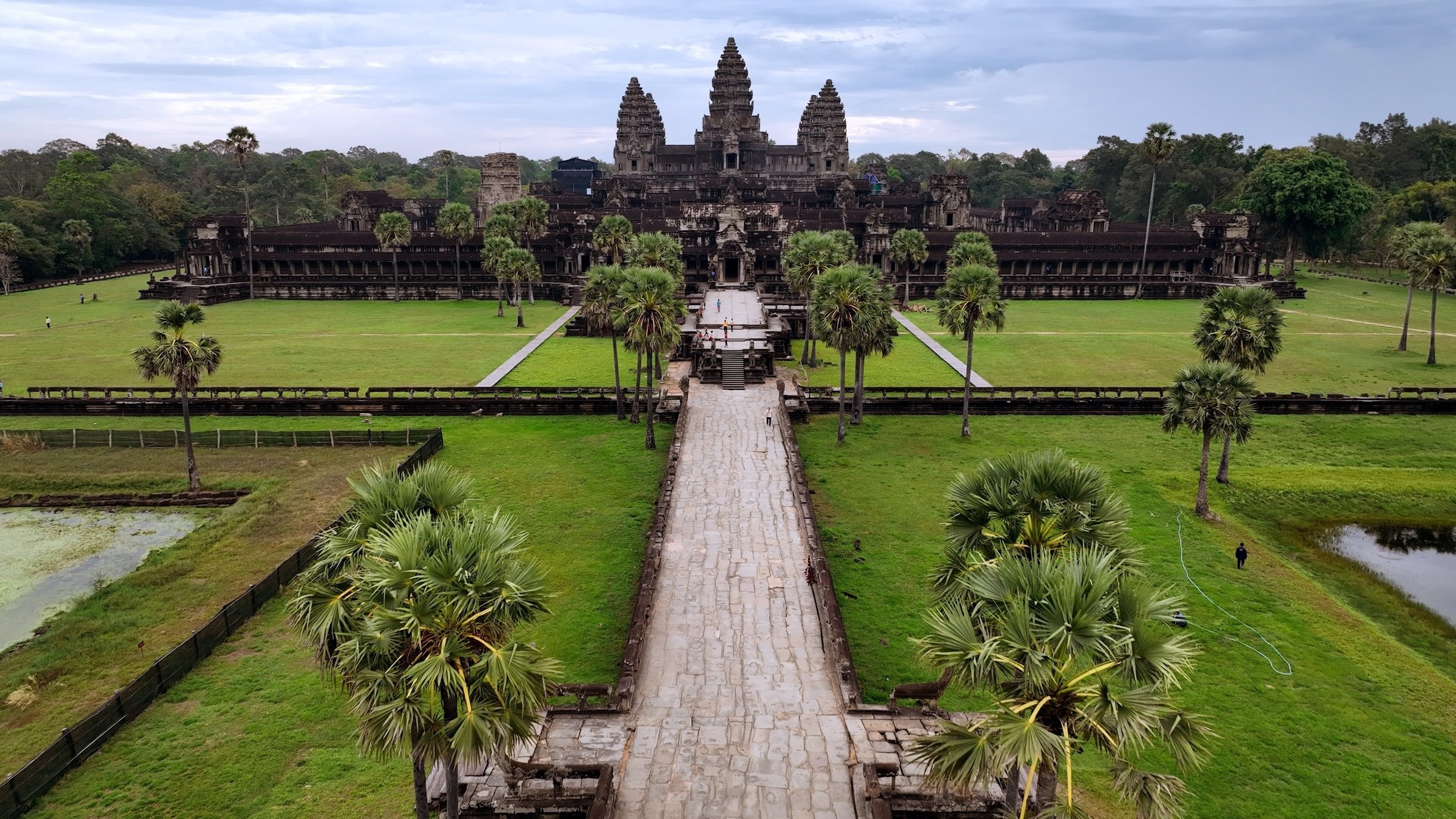 "Kambodscha - Asiens unbekanntes Juwel": Blick von schräg oben auf die Tempelanlage von Angkor Watt mit einer großen Grünfläche vor dem Haupttor. Ein breiter Weg gesäumt von einigen wenigen Palmen führt direkt auf die Anlage zu.