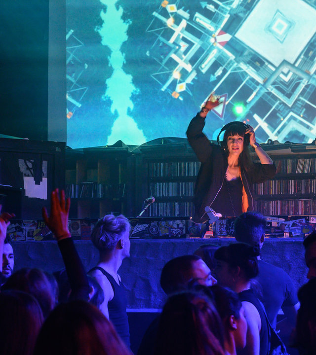 "SOKO Stuttgart - Der letzte Beat": Die Clubbesitzerin Talida Hart (Oona von Maydell) steht etwas erhöht auf dem DJ-Pult im Club "Märzhasen" und legt Musik auf. Mit der einen Hand hält sie die Kopfhörer auf ihrem Kopf fest, die andere Hand hebt sie zum Rhythmus der Musik. Auf der Tanzfläche vor ihr tanzen die Besucher des Clubs (Komparsen). Der hohe Raum ist in blaues Licht getaucht.