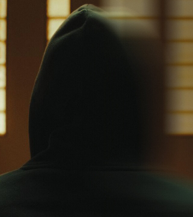 "Putins Krieger - Ein russischer Frontoffizier packt aus": Andrej trägt einen schwarzem Hoodie und hat die Kapuze auf dem Kopf. Er steht in einem Raum mit Glasbausteinen und ist unkenntlich, verschwommen dargestellt.