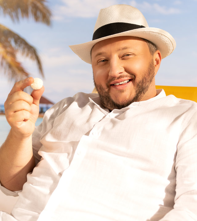 "ZDFzeit: Die Tricks von Haribo, Ferrero & Co. - ZDFbesseresser: Sebastian Lege deckt auf": Sebastian Lege sitzt im Liegestuhl am Strand. In seiner rechten Hand hält er ein Raffaello-Bällchen.