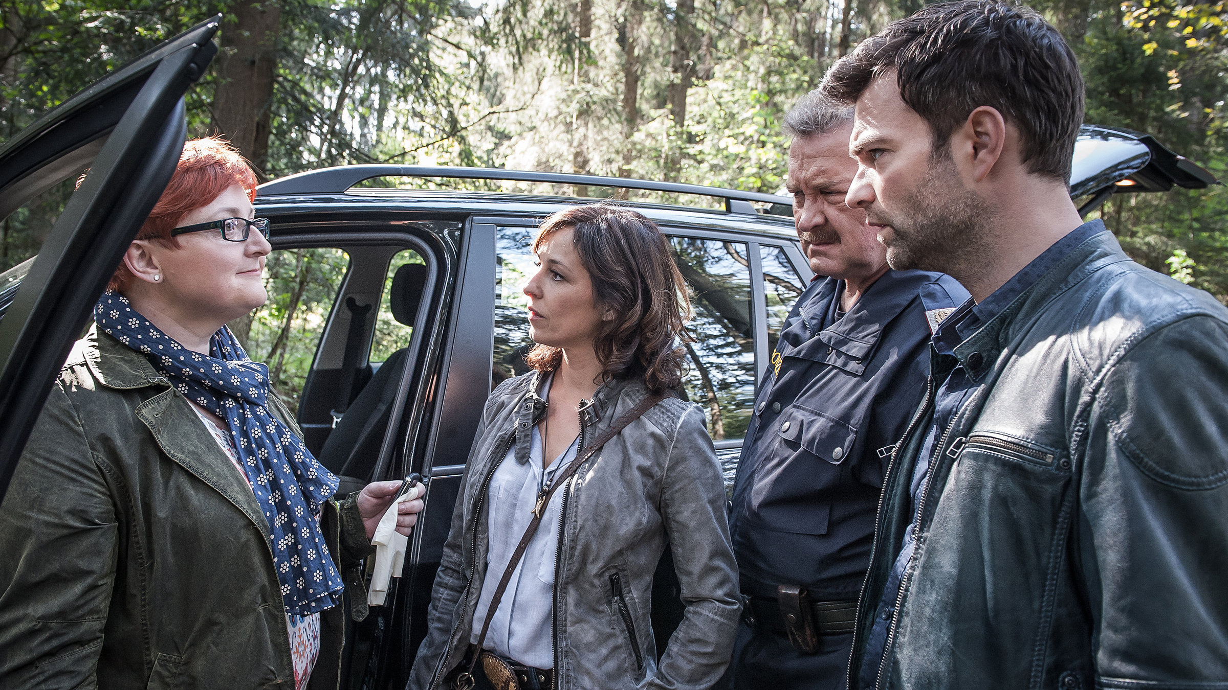 "SOKO Kitzbühel - Vermächtnis": Stefanie (Veronika Polly), Nina (Julia Cencig), Kroisi (Ferry Öllinger) und Lukas (Jakob Seeböck) stehen vor einer offenen Autotür im Wald. Die drei schauen Stefanie überrascht an.
