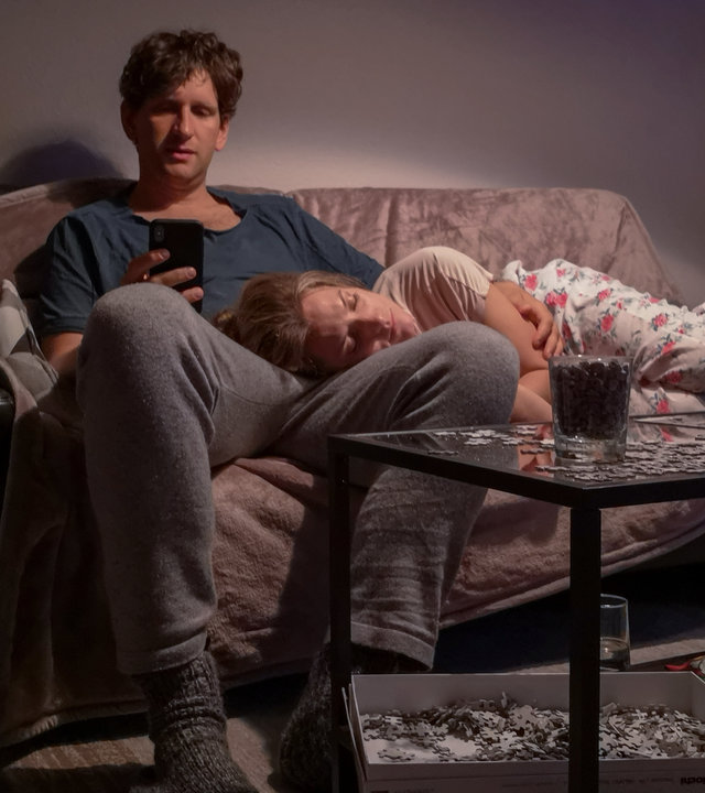 "Liebe. Jetzt! - Der Kuss":  Heiner (Sebastian Schwarz) sitzt mit seinem Handy auf dem Sofa. Seine Frau Meike (Marie Burchard) liegt schlafend auf seinem Schoß.