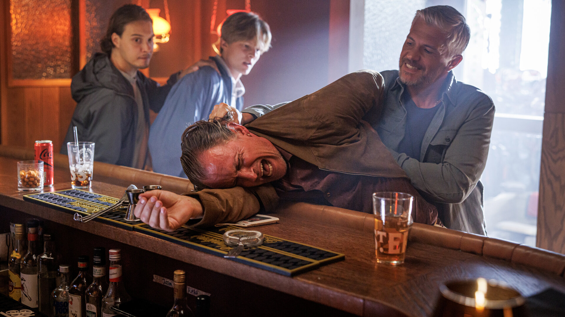 "Mord im Mittsommer - Rakel": Alexander (Nicolai Cleve Broch) packt Ronny (Peter Gröning) am Kragen und drückt ihn in einer Kneipe auf den Tresen. Ronny schreit auf. Im Hintergrund ist Welpe (Julius Fleischanderl) mit einem jungen Mann (Komparse) zu sehen. Er hält ihn an der Schulter fest. Durch die Eingangstür zum Gastraum fällt Tageslicht ein.