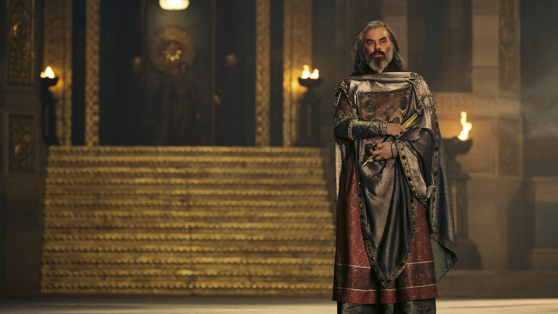 "Der Herr der Ringe: Die Ringe der Macht - Hallen aus Stein": Ar-Pharazôn (Trystan Gravelle) steht in einem großen Saal vor einer goldenen Treppe. Im Hintergrund sind mehrere Feuerschalen und in der Unschärfe weitere Personen zu erkennen.