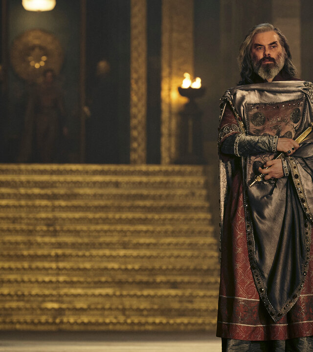 "Der Herr der Ringe: Die Ringe der Macht - Hallen aus Stein": Ar-Pharazôn (Trystan Gravelle) steht in einem großen Saal vor einer goldenen Treppe. Im Hintergrund sind mehrere Feuerschalen und in der Unschärfe weitere Personen zu erkennen.