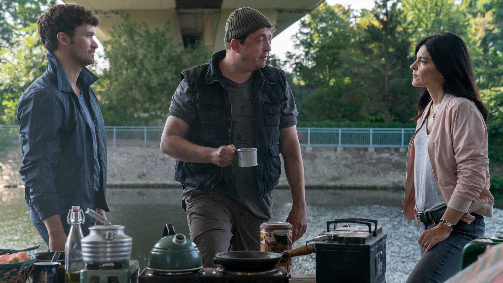 "Letzte Spur Berlin - Verbaut": Kosta (Matthias Lamp) steht mit einer Kaffeetasse in der Hand mit Mina (Jasmin Tabatabei) und Alexander (Aleksandar Radenković) am Rand eines Flusses unter einer Brücke. Sie sprechen miteinander. Vor ihnen ist ein Tisch mit verschiedenen Kochutensilien aufgebaut.