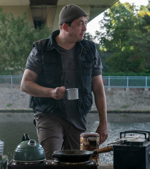 "Letzte Spur Berlin - Verbaut": Kosta (Matthias Lamp) steht mit einer Kaffeetasse in der Hand mit Mina (Jasmin Tabatabei) und Alexander (Aleksandar Radenković) am Rand eines Flusses unter einer Brücke. Sie sprechen miteinander. Vor ihnen ist ein Tisch mit verschiedenen Kochutensilien aufgebaut.