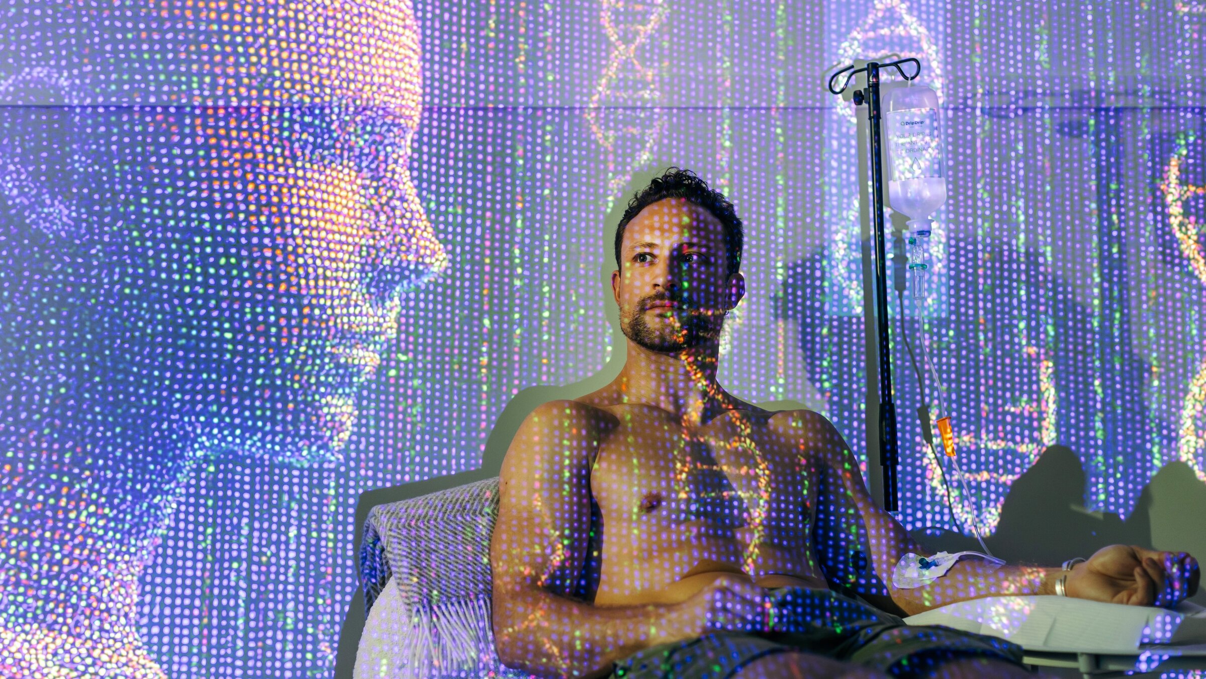 "37°Leben - Longevity": Montage: Ein junger, gut durchtrainierter Mann sitzt mit freiem Oberkörper auf einem Sessel und erhält eine Infusion. Über dem gesamten Bild liegt eine futuristische Grafik mit Doppel-Helix und Lichtpunkten. Ein menschliches Profil aus Lichtpunkten schaut auf Lukas.