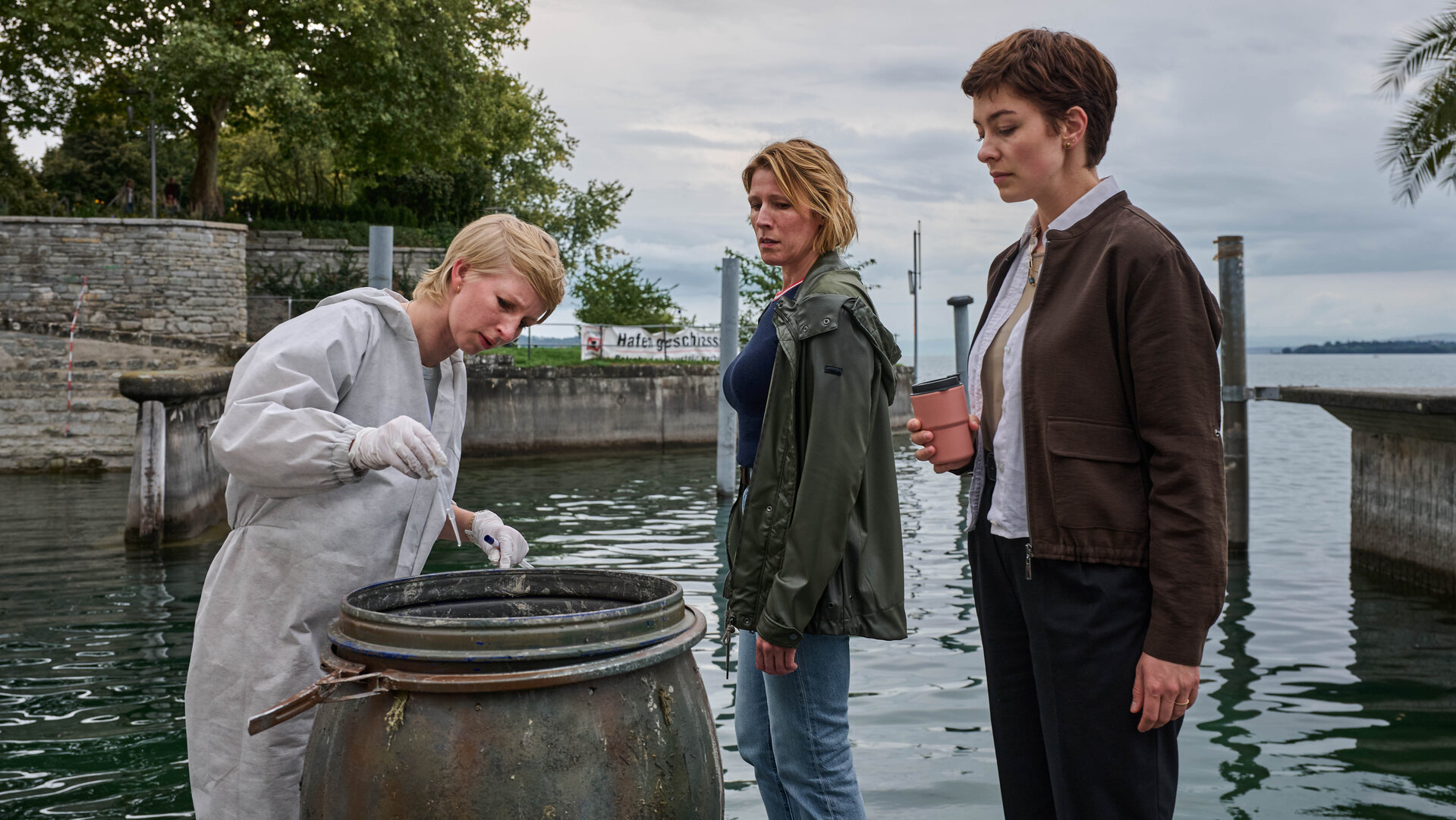 "Mordufer - Kurzes Glück": Sarah Hertweg (Anna Gesa-Raija Lappe), Doro Beitinger (Franziska Weisz) und Chiara Locatelli (Maria Wördemann) stehen am Ufer des Bodensees vor einem offenen Fass. Hertweg trägt einen weißen Overall und weiße Handschuhe, mit der rechten Hand hält sie eine Spritze über das Fass. Beitinger und Locatelli beobachten sie dabei.