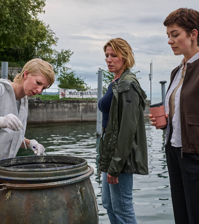 "Mordufer - Kurzes Glück": Sarah Hertweg (Anna Gesa-Raija Lappe), Doro Beitinger (Franziska Weisz) und Chiara Locatelli (Maria Wördemann) stehen am Ufer des Bodensees vor einem offenen Fass. Hertweg trägt einen weißen Overall und weiße Handschuhe, mit der rechten Hand hält sie eine Spritze über das Fass. Beitinger und Locatelli beobachten sie dabei.
