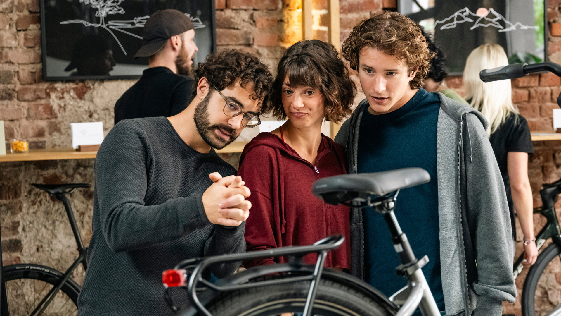 "SOKO Köln -  Bestes Leben": Farsad Reza (Giovanni Funiati), Hanna (Sophie Schlüter) und Tom (Benedikt Kalcher) stehen dicht beieinander in einem Fahrradladen. Sie begutachten aufmerksam ein Fahrrad, das vor ihnen steht. Im Hintergrund hängen zwei gerahmte Bilder an einer Backsteinwand, vor der sich zwei weitere Personen (Komparsen) befinden.