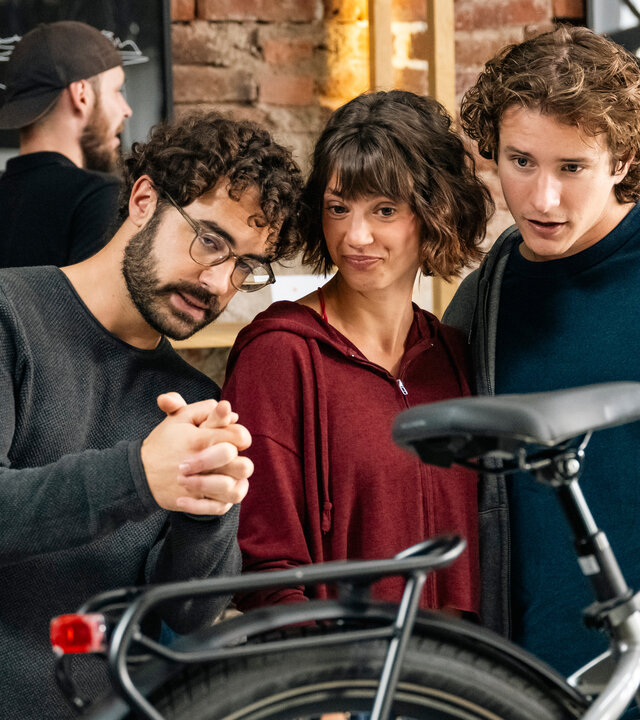"SOKO Köln -  Bestes Leben": Farsad Reza (Giovanni Funiati), Hanna (Sophie Schlüter) und Tom (Benedikt Kalcher) stehen dicht beieinander in einem Fahrradladen. Sie begutachten aufmerksam ein Fahrrad, das vor ihnen steht. Im Hintergrund hängen zwei gerahmte Bilder an einer Backsteinwand, vor der sich zwei weitere Personen (Komparsen) befinden.