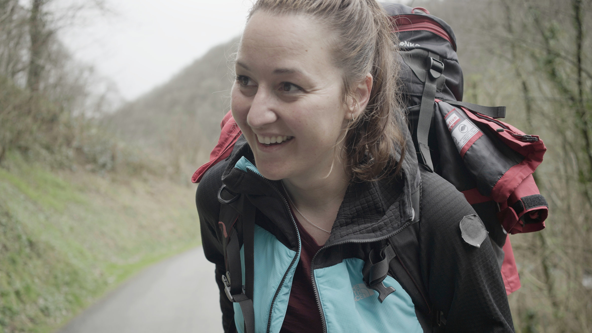 "Mein härtester Weg - Pilgern nach Santiago de Compostela": Aniko in Outdoor-Bekleidung steht auf einer Straße in hügliger Landschaft bei schlechten Wetter und lächelt.