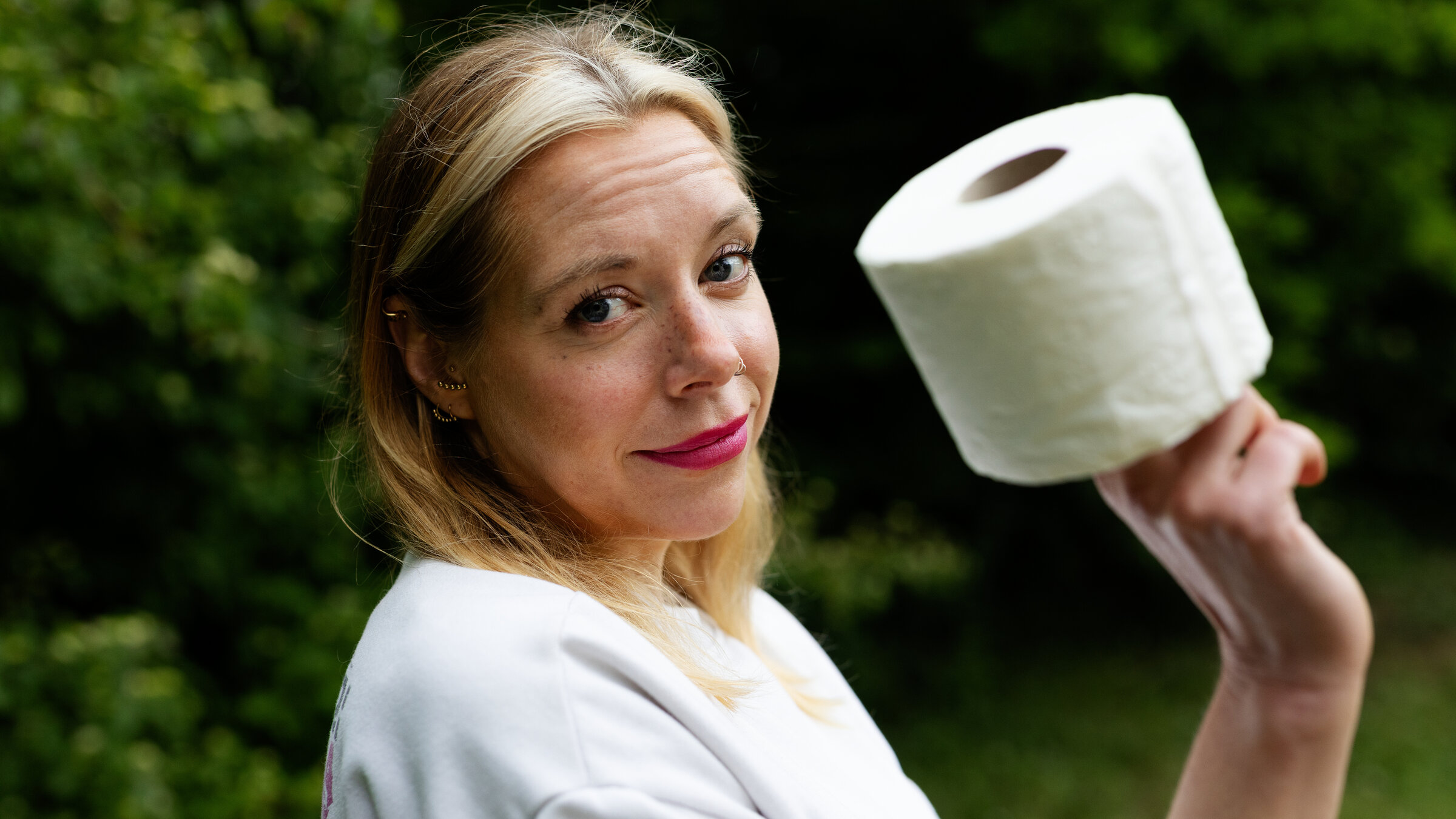 "37°Leben: Müssen müssen - jung und inkontinent": Birgit, eine 40-jährige Frau mit langen blonden Haaren und einem Nasenpiercing, steht seitlich zur Kamera vor grünen Büschen. In der rechten Hand hält sie eine Rolle Toilettenpapier. Ihr Blick ist skeptisch bis fragend, dabei schmunzelt sie ein bisschen.