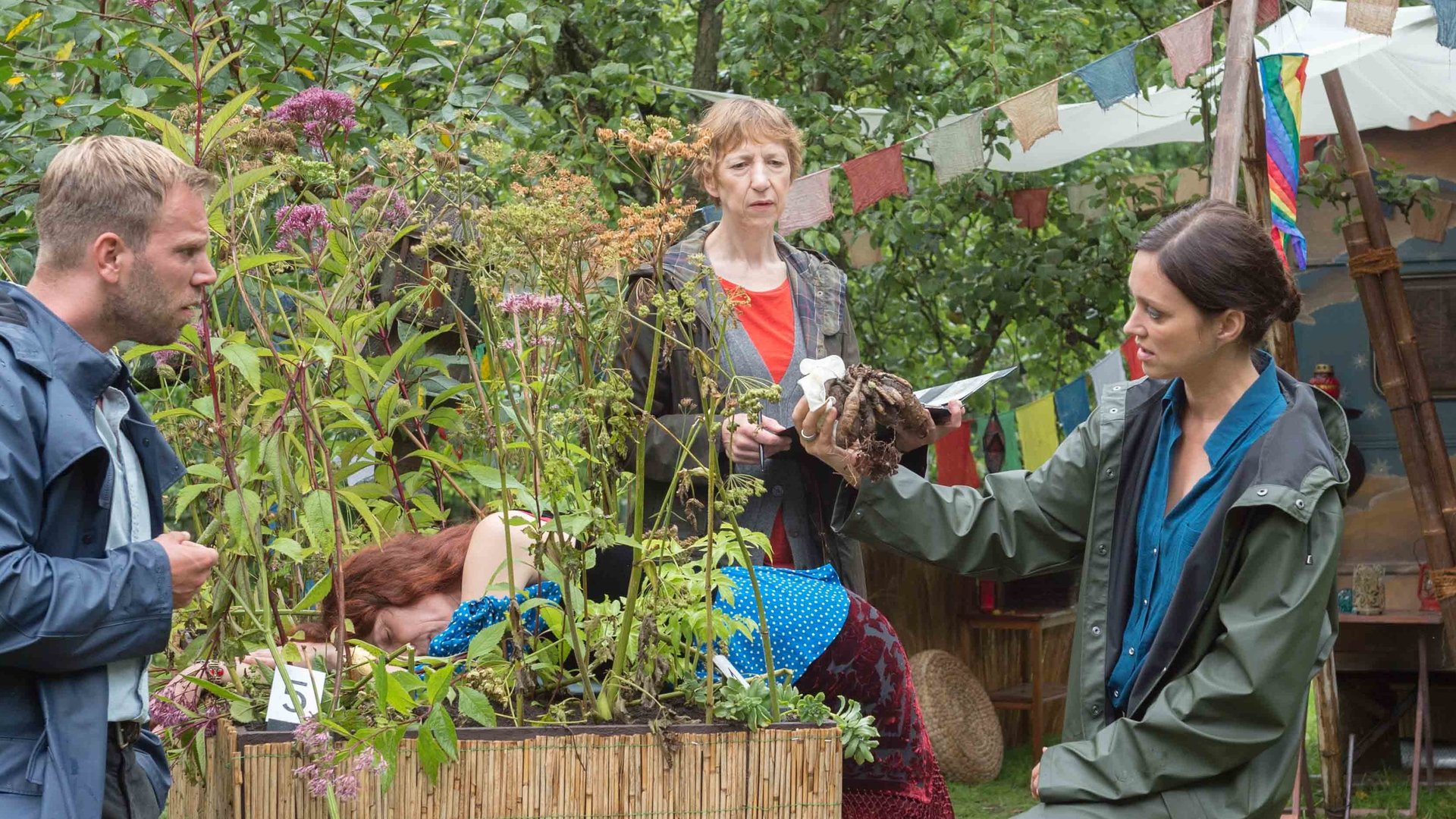 "SOKO Wismar – Einsam“: Die Kommissare Karoline Joost (Nike Fuhrmann) und Lars Pöhlmann (Dominic Boeer) stehen gemeinsam mit Rechtsmedizinerin Dr. Helene Sturbeck (Katharina Blaschke) um ein Kräuterhochbeet, worin Viola Zankovas (Cornelia Horn) mit dem Körper vorne übergebeugt liegt.