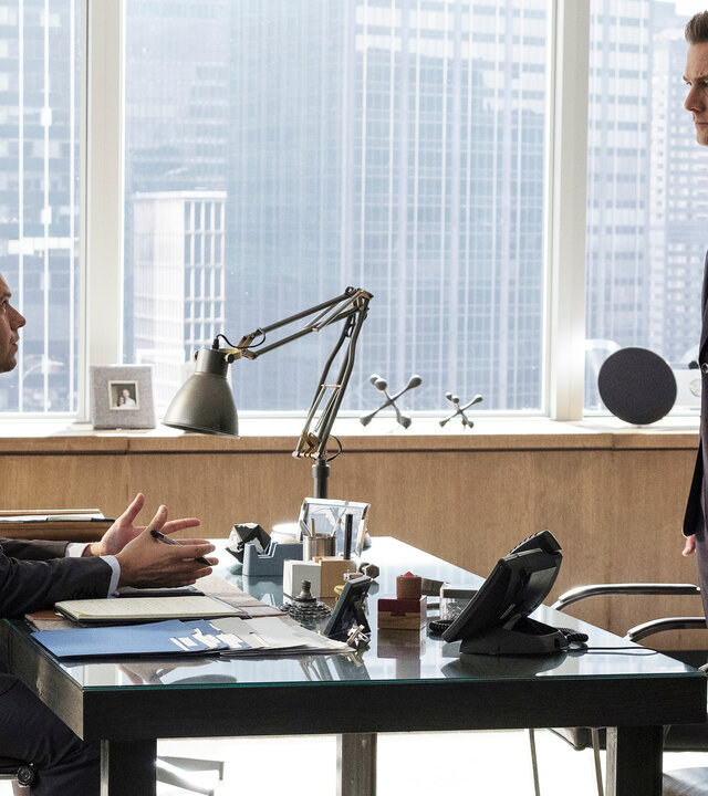 "Suits - Neuordnung": Mike (Patrick J. Adams) sitzt am Schreibtisch, beide Hände auf dem Tisch abgelegt. Er gestikuliert dabei und schaut den vor ihm stehenden Harvey (Gabriel Macht) eindringlich an. Durch die Fensterfront sind im Hintergrund Hochhäuser New Yorks zu erkennen.
