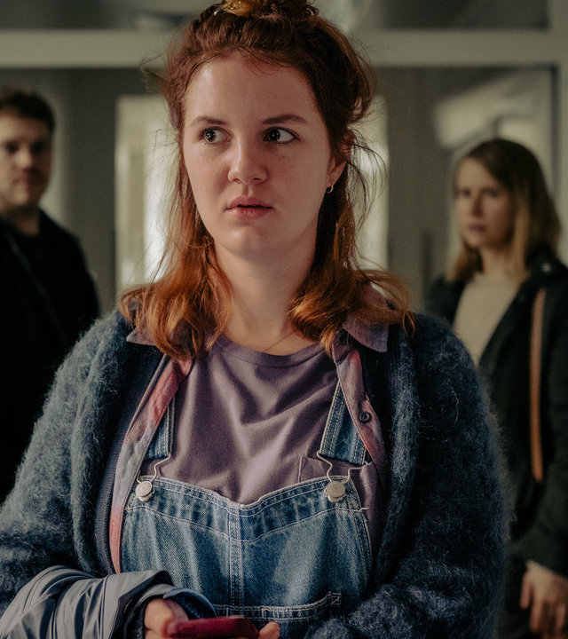 "Another Monday - Schlaflos": Sophie (Alina Stiegler) blickt in die Ferne. Sie trägt eine Latzhose und einen Wollpullover. Im Hintergrund sind Moritz Becker (Ben Münchow) und Freya (Susanne Bormann) zu sehen.