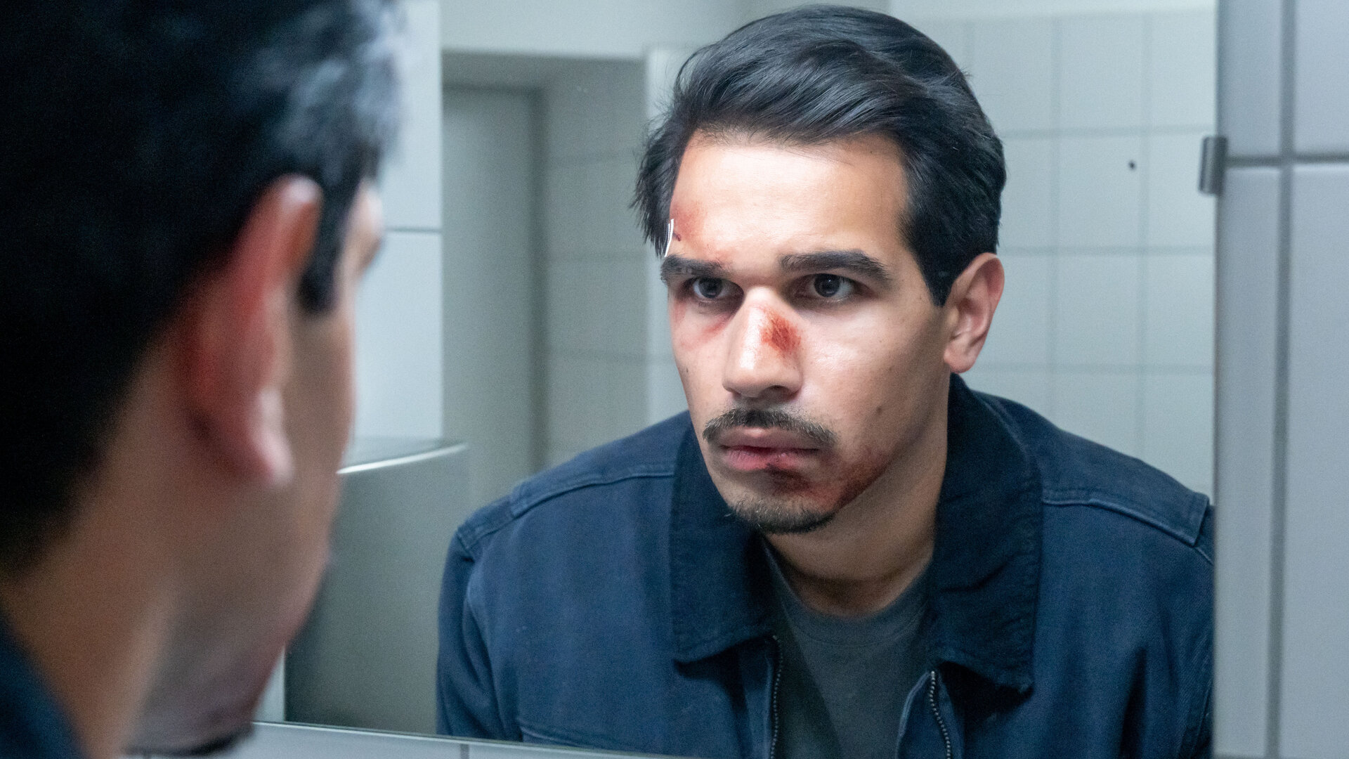 "SOKO Potsdam - Alle hassen Hugo": In einem grell ausgeleuchteten Waschraum steht Ermittler Samir Amari (Skandar Amini) dicht vor einem Spiegel. Sein Gesicht ist von frischen Wunden gezeichnet – die Nase blutig, eine Schramme an der Stirn. Starr blickt er sich selbst in die Augen.