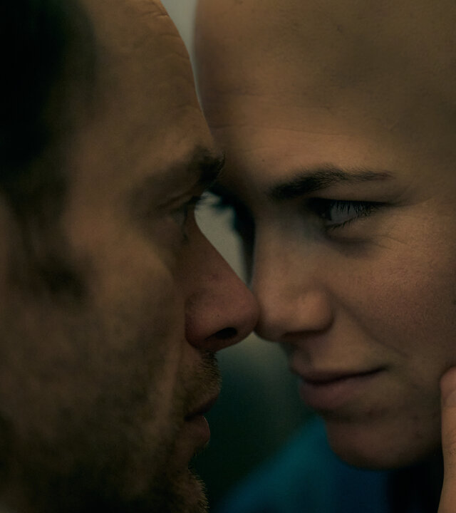 "Don't Fall, Dance - Wenn sich eine Tür schließt, öffnet sich eine andere": Emma (Hanna van Vliet) trägt als Folge der Chemotherapie eine Glatze. Alex (Thomas Ryckewaert) berührt ihr Gesicht mit einer Hand. Sie sitzen sich in unmittelbarer Nähe gegenüber. Ihre Nasen berühren sich.