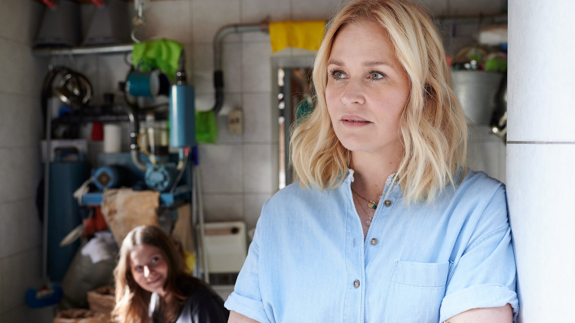 "Lena Lorenz - Harter Bruch": Im Wirtschaftsraum eines Bauernhofes lehnt Lena Lorenz (Judith Hoersch) mit verschränkten Armen an einer Wand. Im Hintergrund sitzt Josi Brenner (Muriel Bielenberg) und schaut lächelnd zu ihr hinüber.