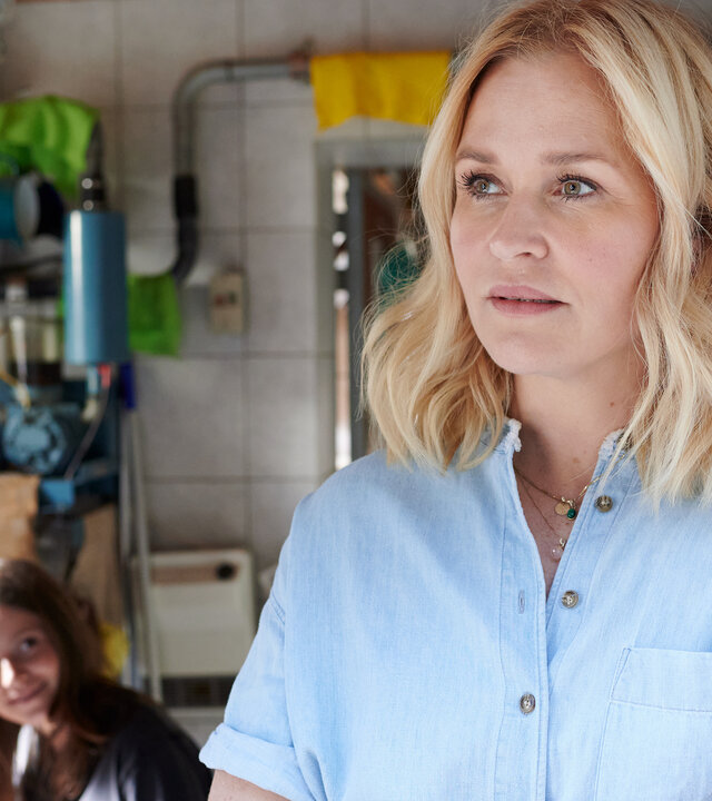 "Lena Lorenz - Harter Bruch": Im Wirtschaftsraum eines Bauernhofes lehnt Lena Lorenz (Judith Hoersch) mit verschränkten Armen an einer Wand. Im Hintergrund sitzt Josi Brenner (Muriel Bielenberg) und schaut lächelnd zu ihr hinüber.