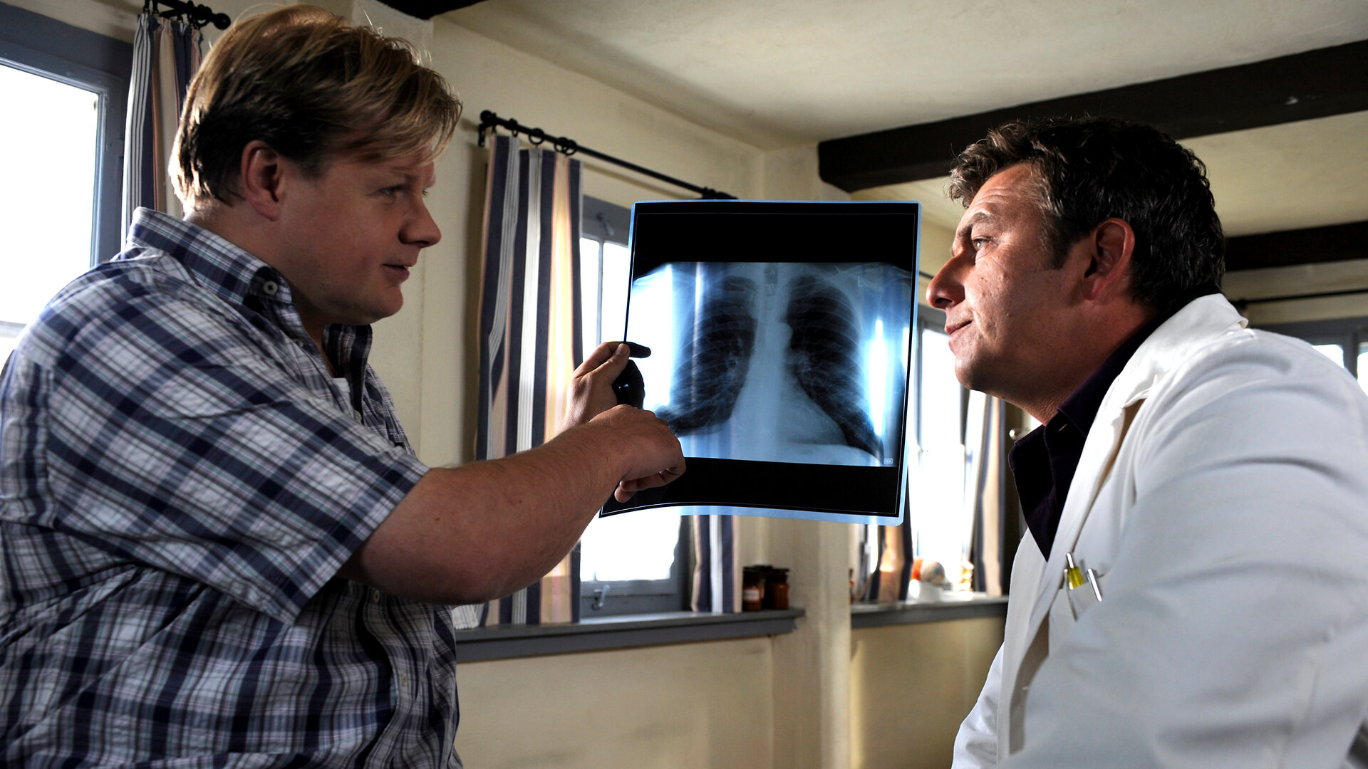 "Der Bergdoktor: Wunderheilung": In der Arztpraxis: Auf der Behandlungsliege sitzt Markus Leitner (Michael A. Grimm) und zeigt auf ein Röntgenbild in seiner Hand. Dabei blickt er wissend Dr. Martin Gruber (Hans Sigl) an, der mit besorgtem Blick vor ihm auf einem Stuhl sitzt.