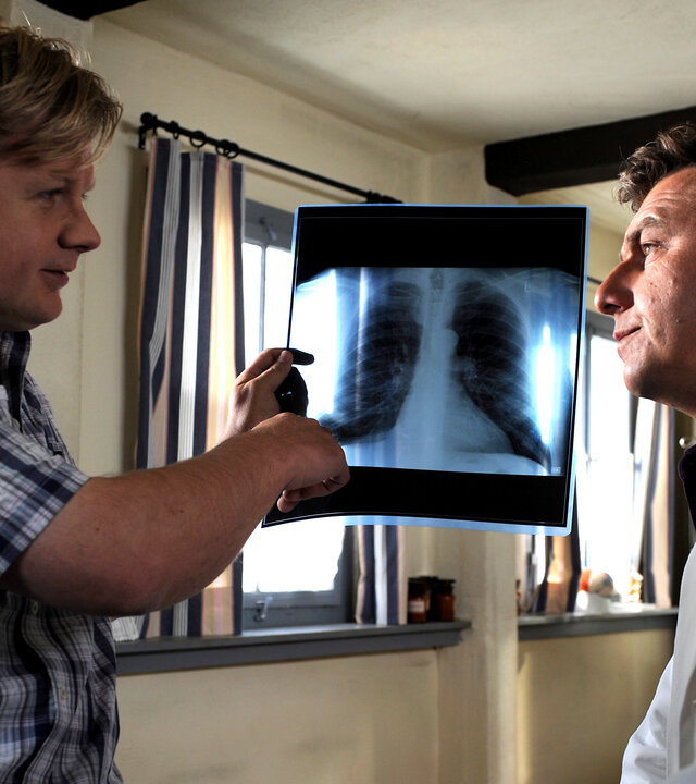 "Der Bergdoktor: Wunderheilung": In der Arztpraxis: Auf der Behandlungsliege sitzt Markus Leitner (Michael A. Grimm) und zeigt auf ein Röntgenbild in seiner Hand. Dabei blickt er wissend Dr. Martin Gruber (Hans Sigl) an, der mit besorgtem Blick vor ihm auf einem Stuhl sitzt.
