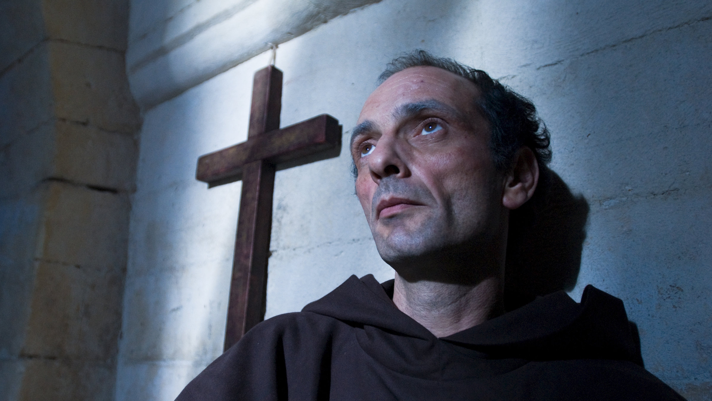 "Die Deutschen, Heinrich und der Papst" - Papst Gregor VII. (Gabriel Spahiu) in Nahaufnahme vor einem Holzkreuz, dass an einer Wand hängt.