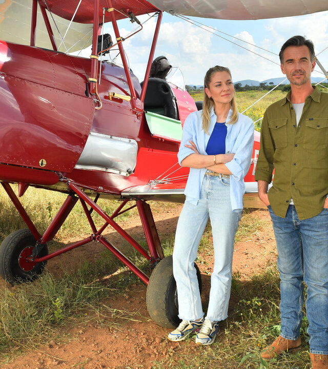 "Das Traumschiff: Afrika: Madikwe": Ein rotes Doppeldeckerflugzeug steht in der Savanne. Sophia Berg (Valentina Pahde) lehnt lässig an einem Flügel und blickt verschmitzt zu Max Parger (Florian Silbereisen). Dieser steht, die Hände in den Hosentaschen, neben ihr und blickt lächelnd in die Ferne.
