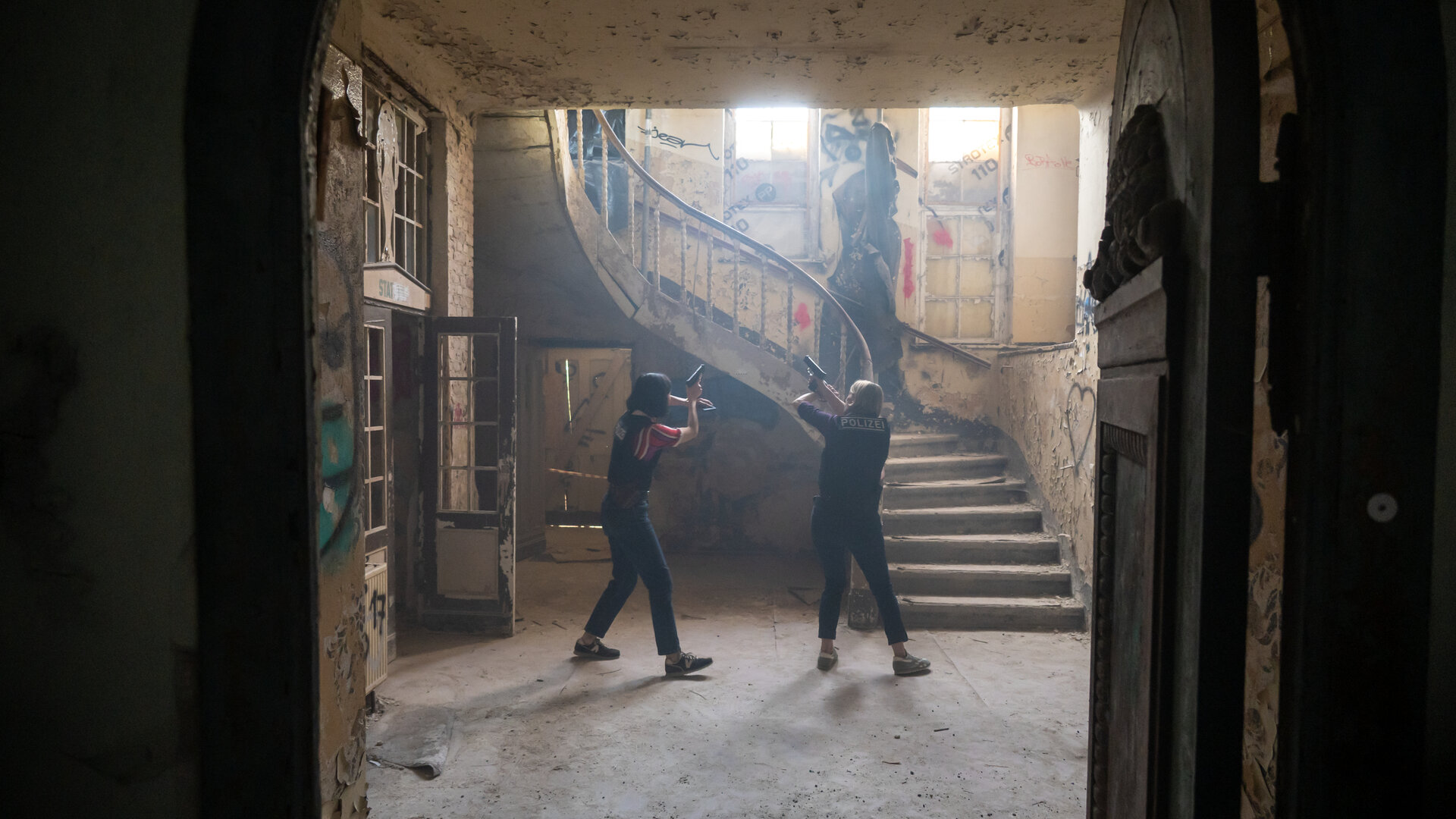 "SOKO Potsdam - Lost": In der Eingangshalle einer stark verfallenen Psychiatriegebäudes stehen die Kommissarinnen Tamara Meurer (Anja Pahl) und Pauline Hobrecht (Agnes Decker) mit gezogenen Dienstwaffen. Sie bewegen sich vorsichtig über den staubigen Boden, die Körper angespannt, die Arme erhoben und auf einen Punkt vor sich gerichtet. Das Licht fällt fahl durch hohe, zerbrochene Fenster und beleuchtet eine bröckelnde Treppe mit abblätterndem Geländer. Wände und Türen sind von Graffiti bedeckt, Putz liegt auf dem Boden.