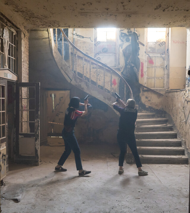 "SOKO Potsdam - Lost": In der Eingangshalle einer stark verfallenen Psychiatriegebäudes stehen die Kommissarinnen Tamara Meurer (Anja Pahl) und Pauline Hobrecht (Agnes Decker) mit gezogenen Dienstwaffen. Sie bewegen sich vorsichtig über den staubigen Boden, die Körper angespannt, die Arme erhoben und auf einen Punkt vor sich gerichtet. Das Licht fällt fahl durch hohe, zerbrochene Fenster und beleuchtet eine bröckelnde Treppe mit abblätterndem Geländer. Wände und Türen sind von Graffiti bedeckt, Putz liegt auf dem Boden.