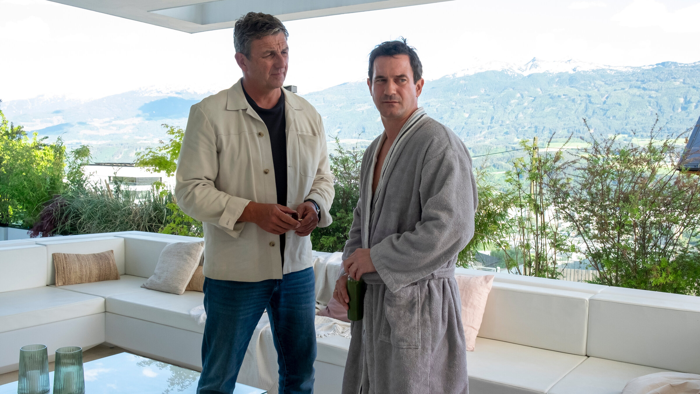 "Der Bergdoktor: Wechselwirkungen": Auf einer Terrasse mit Blick in die Berge: Dr. Martin Gruber (Hans Sigl) steht neben Hendrik Barlan (Sönke Möhring) und blickt ihn stirnrunzelnd an. Hendrik Barlan trägt einen Bademantel und hat sich mit nachdenklichem Blick von Dr. Gruber abgewendet.