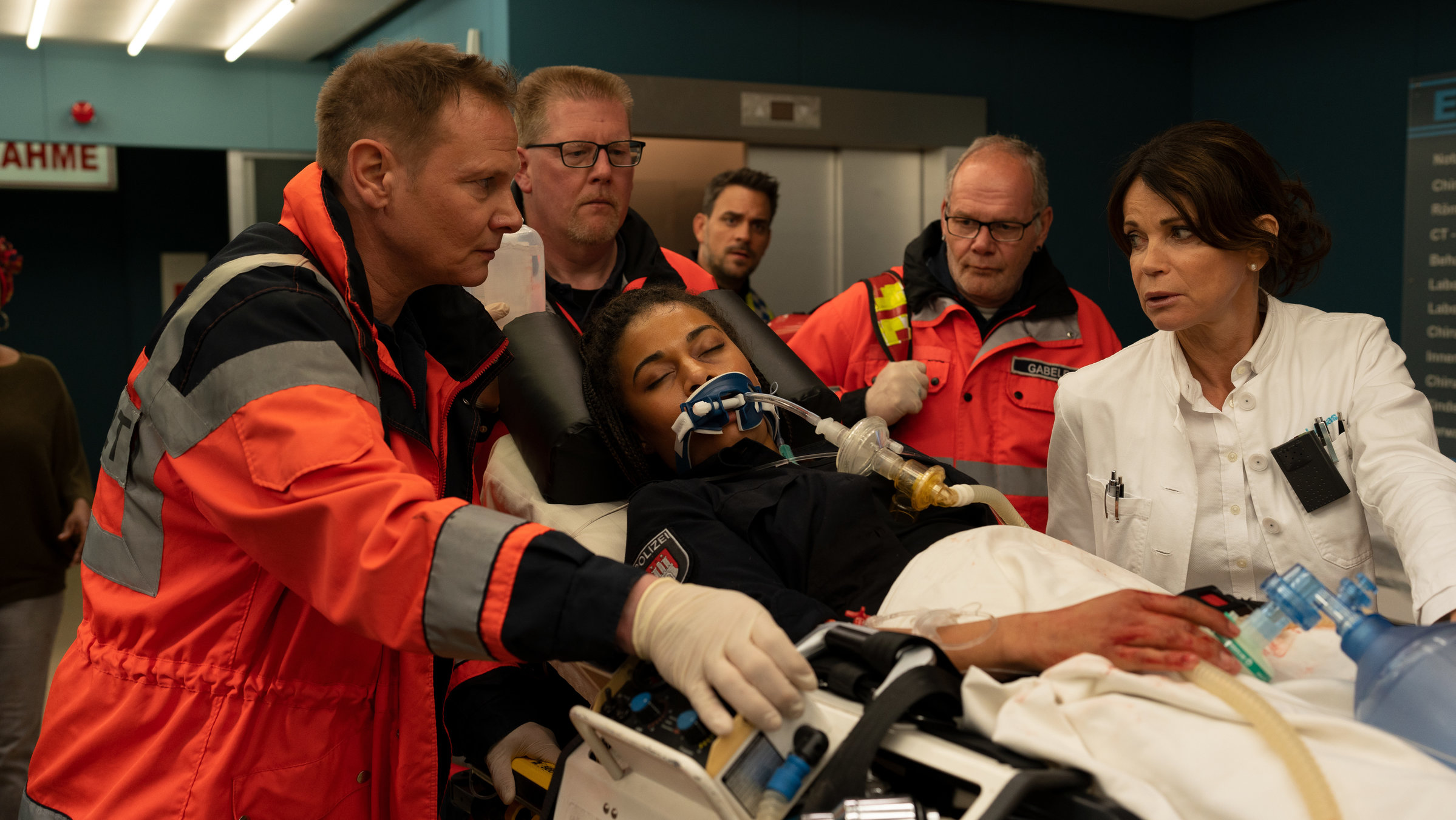 "Notruf Hafenkante - Weil du mir gehörst": In der Notaufnahme des Krankenhauses: Dr. Haase (Fabian Harloff) und Dr. Jonas (Gerit Kling) gehen neben der Patiententrage, auf der die Polizistin Daisy Petersen (Aysha Joy Samuel) liegt. Daisy hat die Augen geschlossen, sie scheint nicht bei Bewusstsein. Sie ist intubiert. Dr. Haase und Dr. Jonas sehen sich besorgt an. Im Hintergrund sind zwei weitere Helfer (Komparsen).