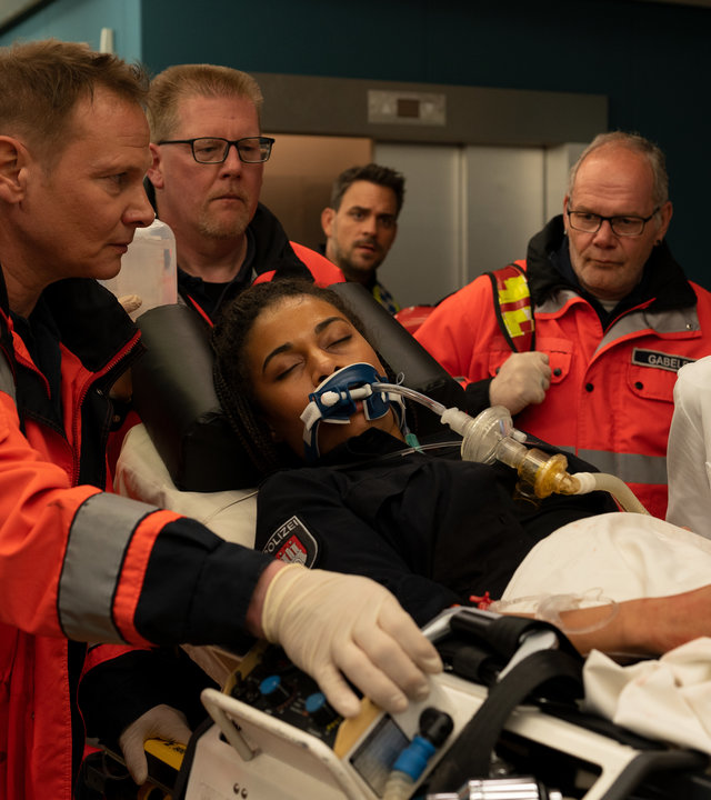 "Notruf Hafenkante - Weil du mir gehörst": In der Notaufnahme des Krankenhauses: Dr. Haase (Fabian Harloff) und Dr. Jonas (Gerit Kling) gehen neben der Patiententrage, auf der die Polizistin Daisy Petersen (Aysha Joy Samuel) liegt. Daisy hat die Augen geschlossen, sie scheint nicht bei Bewusstsein. Sie ist intubiert. Dr. Haase und Dr. Jonas sehen sich besorgt an. Im Hintergrund sind zwei weitere Helfer (Komparsen).