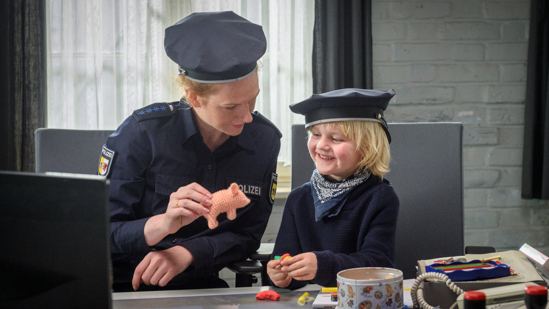 "SOKO Wismar - Wie das Leben spielt": Paula Moorkamp (Stella Hinrichs) sitzt neben Tim Kern (Yvon Sable Moltzen) an ihrem Schreibtisch. Paula spielt mit einem Plastikschwein und Tim ist sichtlich erfreut. Im Vordergrund befinden sich ein Bildschirm, eine Tasse, sowie mehrere Dokumente.