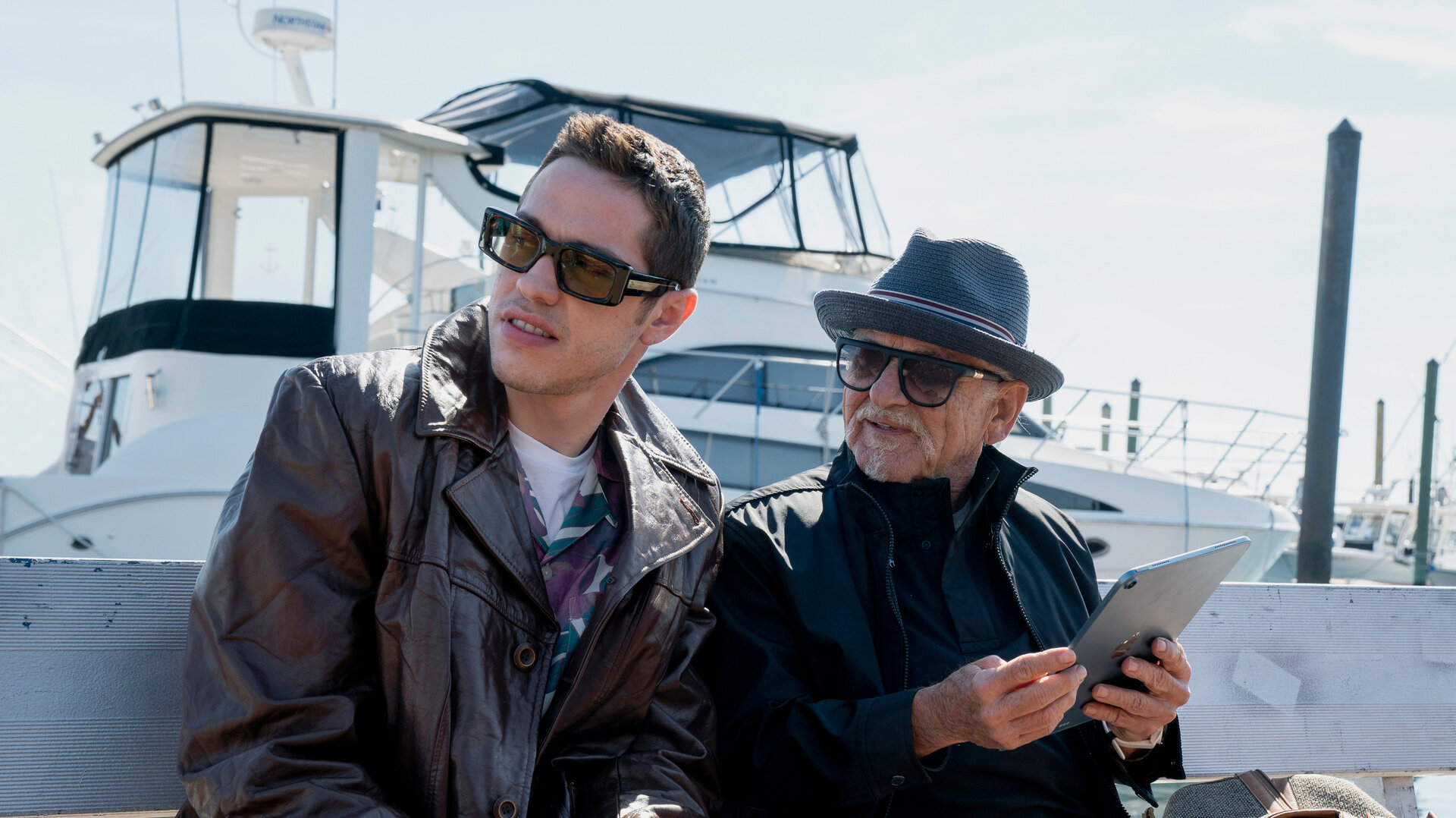 "Bupkis - Das Foto": Pete (Pete Davidson) sitzt cool in Lederjacke und mit Sonnenbrille auf einer Holzbank. Rechts neben ihm der ebenso coole Opa Joe (Joe Pesci). Auch er trägt eine Sonnenbrille und hat seinen Hut lässig ins Gesicht gezogen. Hinter Ihnen liegt eine große Motorjacht im Hafen.