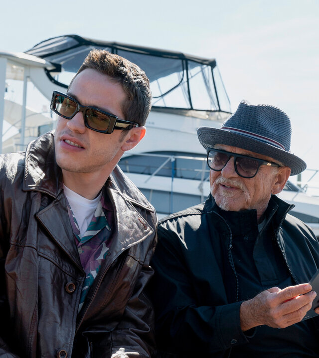 "Bupkis - Das Foto": Pete (Pete Davidson) sitzt cool in Lederjacke und mit Sonnenbrille auf einer Holzbank. Rechts neben ihm der ebenso coole Opa Joe (Joe Pesci). Auch er trägt eine Sonnenbrille und hat seinen Hut lässig ins Gesicht gezogen. Hinter Ihnen liegt eine große Motorjacht im Hafen.