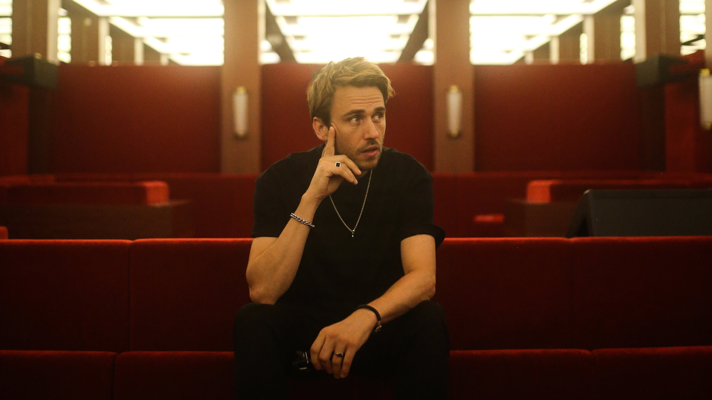 "Clueso: Sommer Tour 2024 - Live von der Berliner Waldbühne" Der Pop-Künstler Clueso sitzt in einem Raum mit hell erleuchteter Decke und mit rotem Samt bezogenen Sitzbänken. Er selbst trägt eine schwarze Hose und T-Shirt und weiße Sneaker. Um die Handgelenke trägt er jeweils ein Armband, um den Hals eine Kette. Er hält eine Hand an sein Kinn und schaut zur Seite.