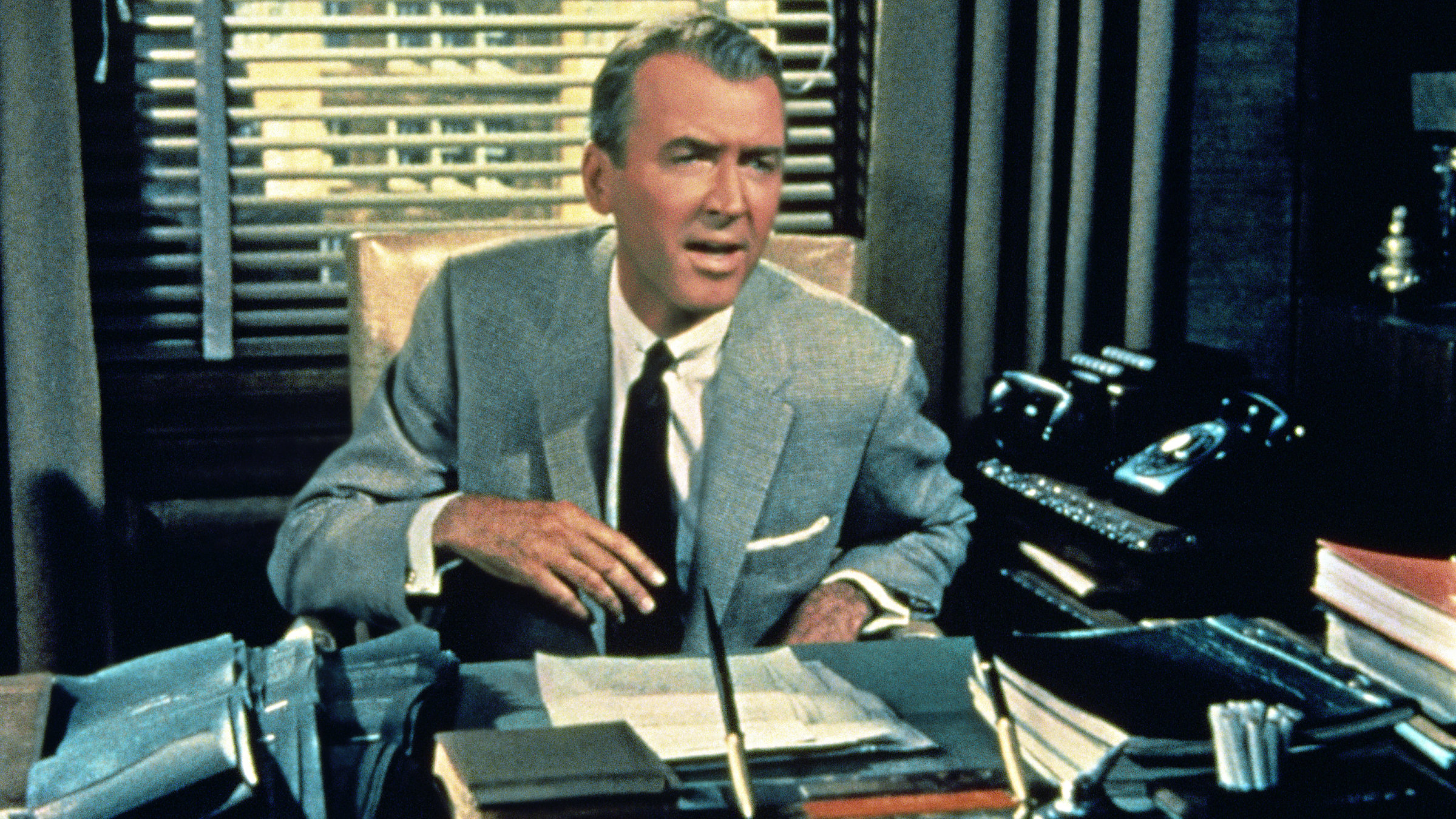 "Meine Braut ist übersinnlich": Hinter einem überfüllten Schreibtisch sitzt der Verleger Shepherd Henderson (James Stewart) in einem hellen Anzug und dunkler Krawatte. Er spricht mit jemandem außerhalb des Bildes und wirkt angespannt und unzufrieden.