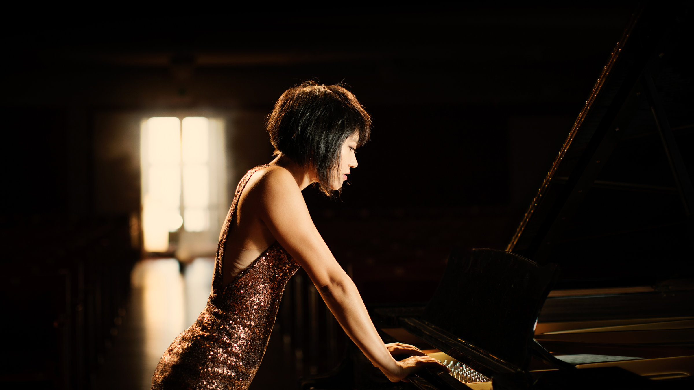 "Sommernachtskonzert Schönbrunn 2019" - Eine Frau steht  in einem schimmernden goldenen Abendkleid an einen Konzertflügel gelehnt und berührt eine Taste.
