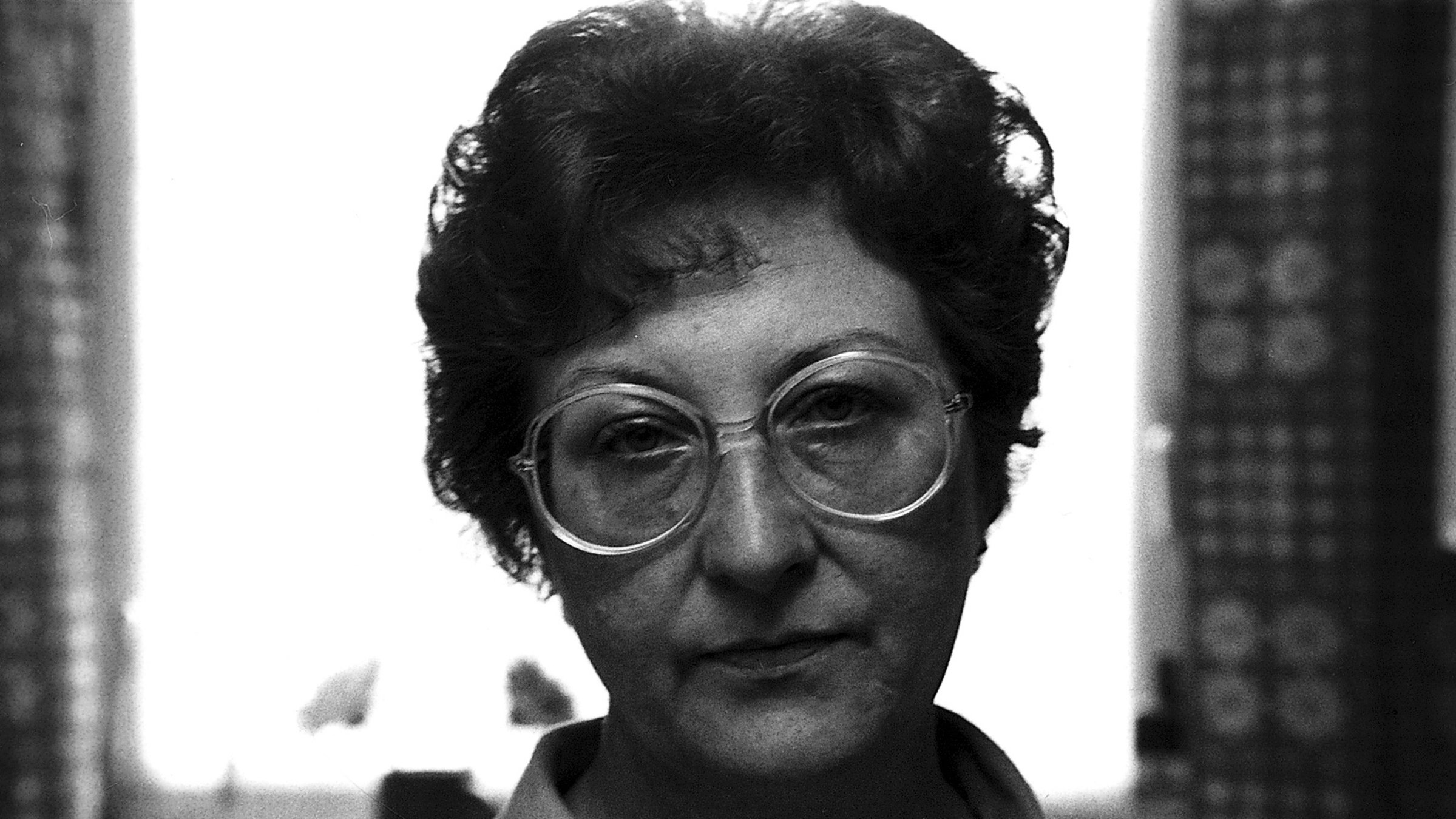 "Von wegen Schicksal": Schwarz-Weiß-Bild: Nahaufnahme von Irene Rakowitz, die direkt in die Kamera blickt. Ihr Gesichtsausdruck ist neutral. Irene Rakowitze hat kurze, gelockte Haare und trägt eine große, runde Brille. Sie steht vor einem Fenster mit gemusterten Vorhängen.