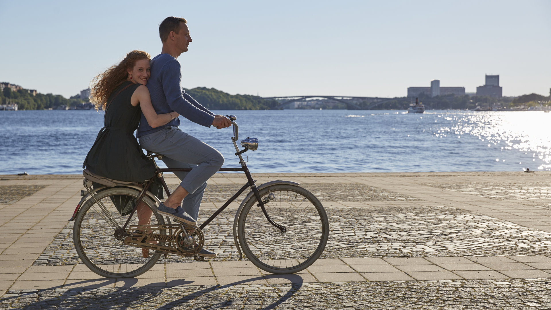 "Inga Lindström: Tanz mit mir": Alban (Mike Hoffmann) fährt Fahrrad und Eva (Sina-Valeska Jung) sitzt hinten auf dem Gepäckträger, sie hält sich an Alban fest - im Hintergrund Wasser und Landschaft.