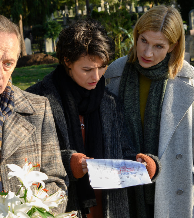 "SOKO Stuttgart – Ruhe in Frieden": Martina Seiffert (Astrid M. Fünderich) und Nele Becker (Nina Siewert) stehen mit dem Unternehmer Amadeus Sandmann (Wilfried Hochholdinger) auf dem Friedhof. Er hält einen Strauß weißer Lilien in der Hand. Es ist ein sonniger Tag. Alle sind in Wintermäntel gekleidet. Nele schaut konzentriert auf ein Dokument, welches sie in der Hand hält. Seiffert schaut Sandmann prüfend von der Seite an, während der bedrückt nach vorne schaut.