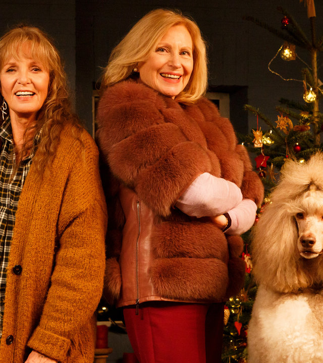 "Mona & Marie": Marie Decker (Ulrike Kriener) und ihre Schwester Mona Berthold (Maren Kroymann) stehen Rücken an Rücken in einem weihnachtlich geschmückten Wohnzimmer. Rechts von ihnen der weiße Königspudel Lucky.