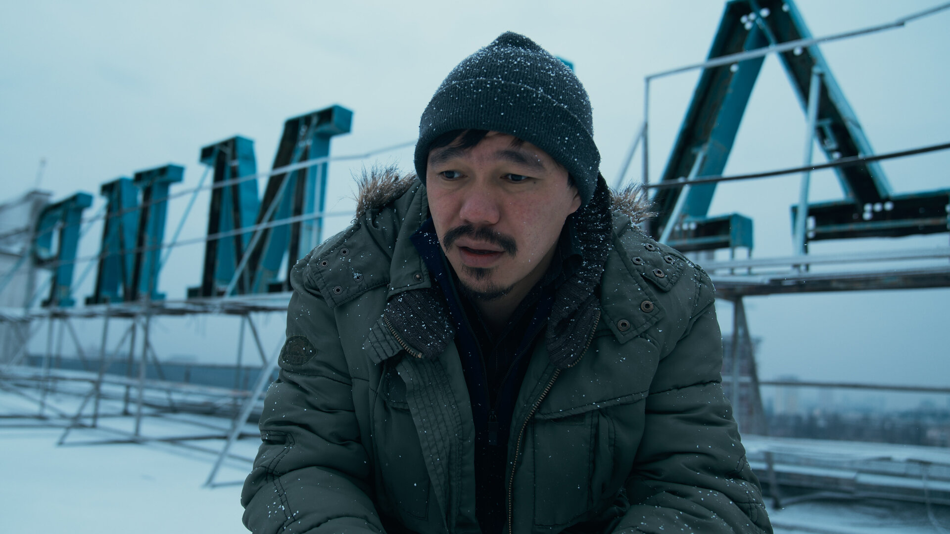 "Infiniti - Turm zu Babel": Isaak (Daniyar Alshinov) kniet auf einem verschneitem Dach und schaut konzentriert in die Ferne. Im Hintergrund ist der kyrillische Schriftzug des Gebäudes zu erkennen.
