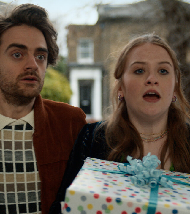 "Extraordinary - Jen vs. Nora: Der ultimative Showdown": Jizzlord (Luke Rollason) und Jen (Máiréad Tyers) stehen vor der Türe. Im Hintergrund stehen Häuser einer schicken Wohngegend. Jen hält ein großes Geschenk, das mit gepunktetem Papier und blauer Schleife eingewickelt ist, in den Händen. Jizzlord schaut erstaunt.