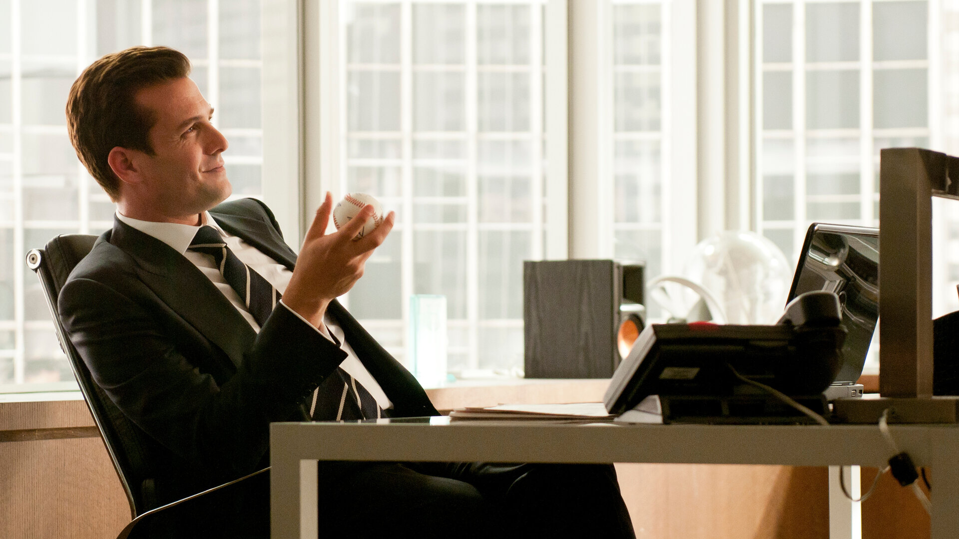 "Suits - Herausforderungen": Harvey (Gabriel Macht) sitzt in einem modernen Büro am Schreibtisch, lächelt leicht und hält einen Baseball in der Hand. Im Hintergrund sind hohe Fensterfronten sehen.