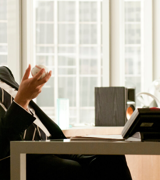 "Suits - Herausforderungen": Harvey (Gabriel Macht) sitzt in einem modernen Büro am Schreibtisch, lächelt leicht und hält einen Baseball in der Hand. Im Hintergrund sind hohe Fensterfronten sehen.