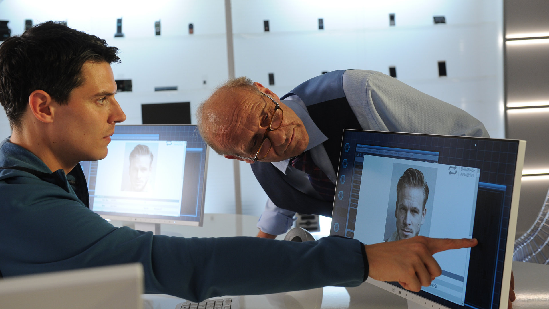 "SOKO Stuttgart - Schuldig": Rico Sander (Benjamin Strecker, l.) sitzt an seinem Schreibtisch und zeigt mit dem rechten Zeigefinger auf den Monitor vor sich. Michael Kaiser (Karl Kranzkowski, r.) beugt sich über den Tisch, um einen besseren Blick auf den Bildschirm zu bekommen, auf dem ein Foto von Julius Weber zu sehen ist.